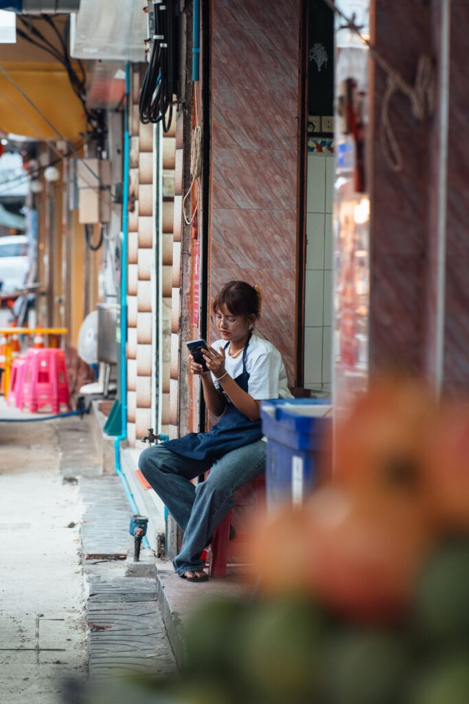 Blumen im vordergrund und es sitzt eine junge thai dame auf der Haustürtreppe und sieht in ihr smartphone fotografiert mit einer langen brennweite von stefan sperl