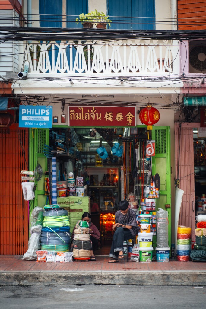 kleiner laden in bangkok chinatwon. zwei personen sitzen davor auf dem boden und sind vertieft in ihre handys fotografiert von stefan sperl mit einer langen Brennweite